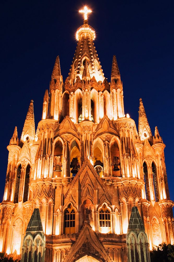Church building with gothic architecture and multiple stone spires illuminated at night with an orange glow and a large cross, Guanajuato, Mexico.