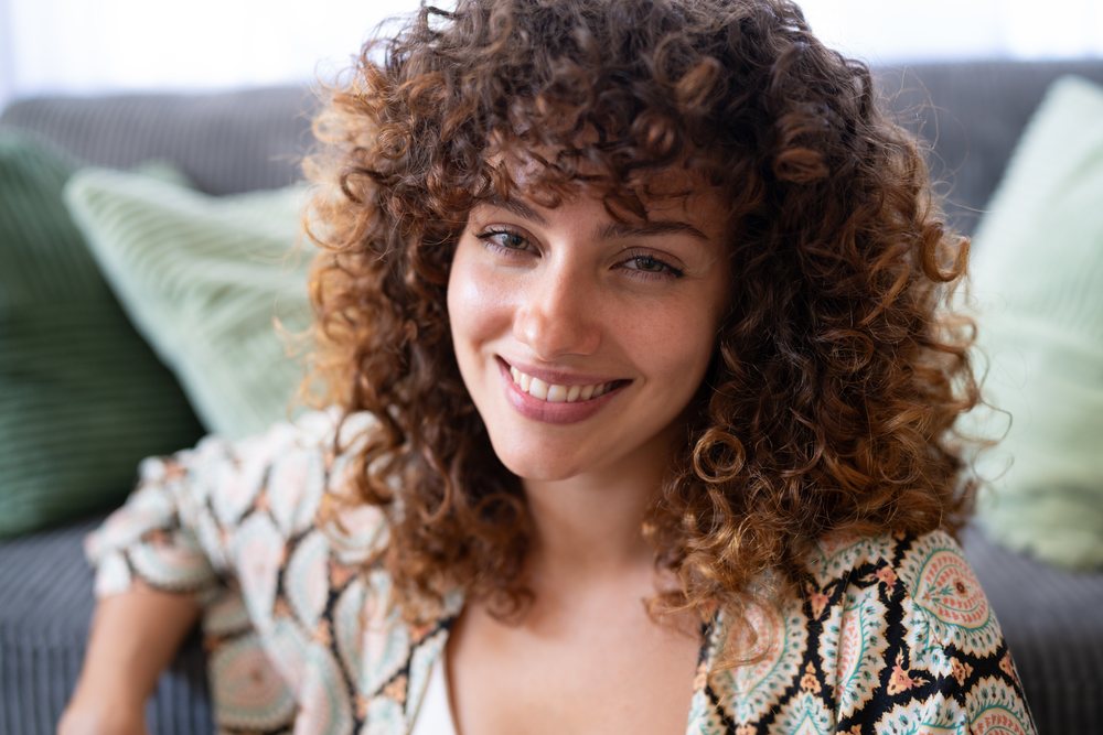 Smiling woman with curly hair sitting on a couch with green cushions in a home living room, Spain.