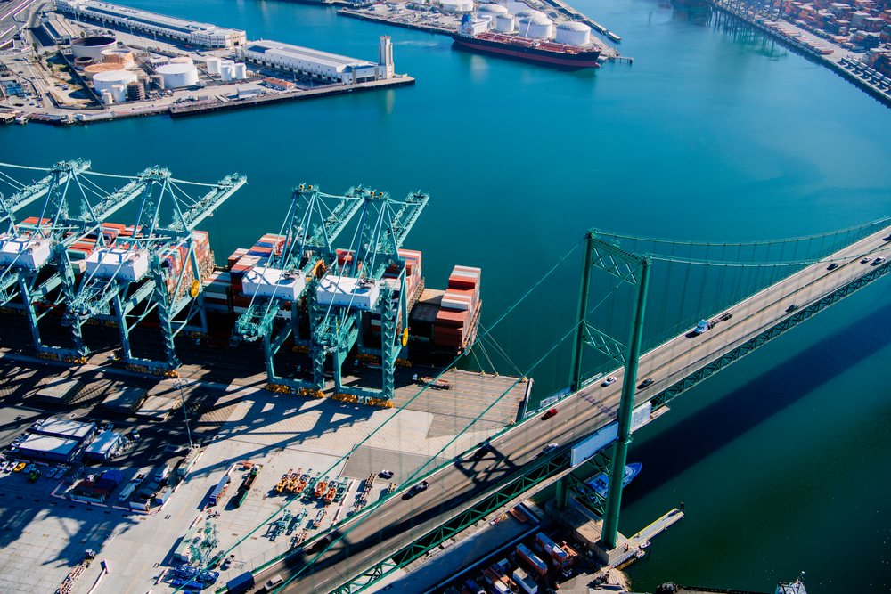 Cargo cranes loading shipping containers at an industrial port with a bridge crossing the water and a cargo ship docked nearby, Port of Long Beach, CA, USA.