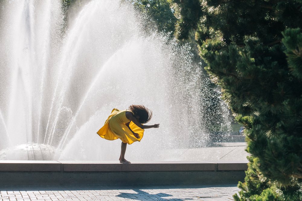 Woman dancing barefoot outdoors in a yellow dress by a large fountain with trees nearby, USA.