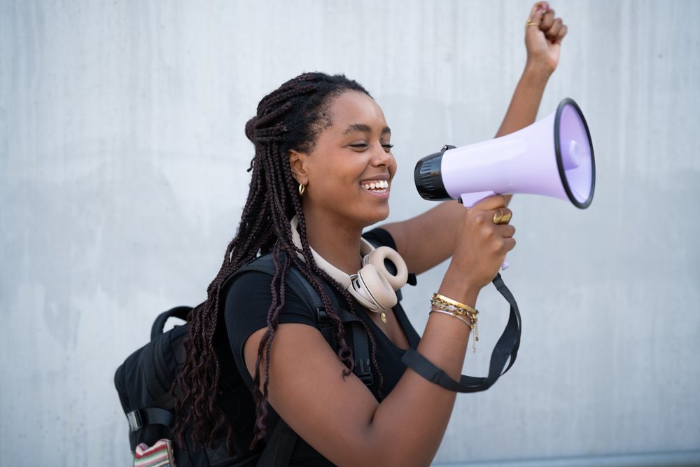 A young woman with braided hair holds a megaphone and shouts happily in an urban setting, Santiago De Compostela, Spain.