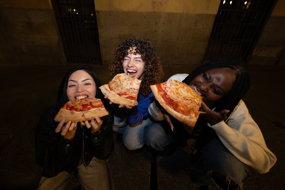 Three women happily eating cheese pizza slices together outdoors at night in an urban setting, Madrid, Spain.