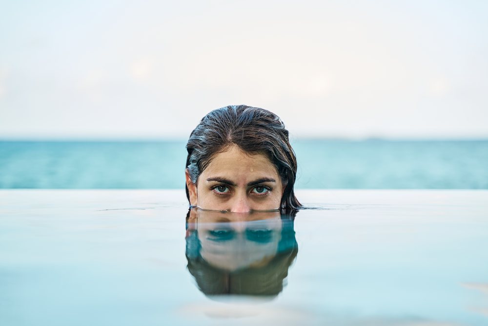 A white woman partially submerged in an outdoor pool with ocean view