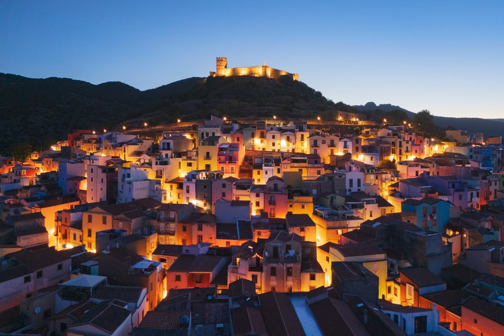 Illuminated hilltop fortress overlooks the densely packed Old Town of Bosa at Dawn with mountain backdrop, Travel Images, Sardinia, Italy.