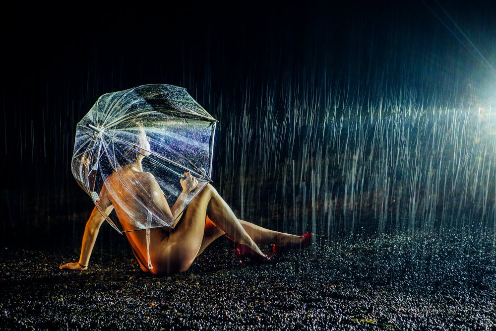 Nude woman wearing red high-heeled shoes sits on wet black gravel under a transparent umbrella in heavy rain at night, cosplay, nudity, freedom, expression, USA.