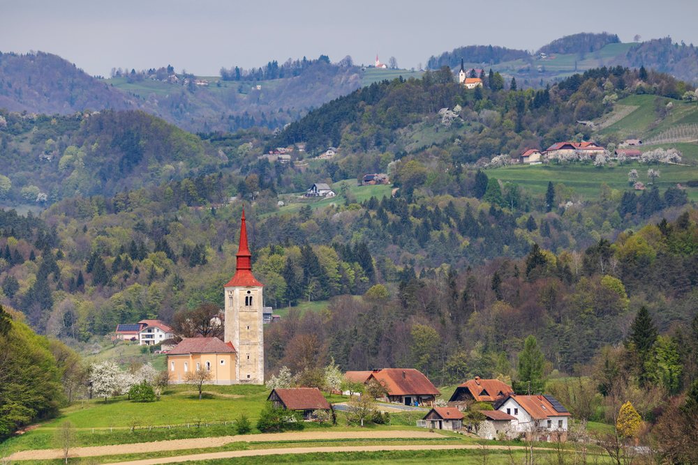 Church with red steeple and multiple red-roofed houses on grassy hill, landscape, cityscape, Mokronog, Slovenia.