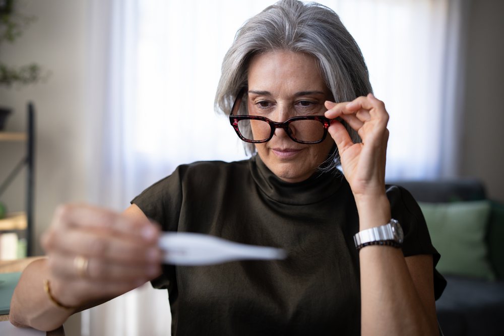 Mature woman with gray hair and glasses reads a thermometer indoors at home, monitoring her temperature, Spain.