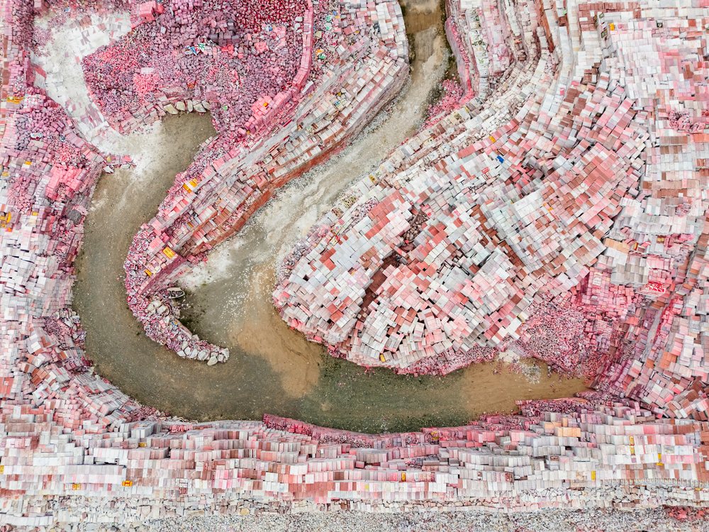 Aerial view of stacked pink and light gray stone blocks and a curved muddy riverbed flowing through a rocky quarry in Kham Tibet, China, Xinzhaia, Qinghai Province, People's Republic of China.