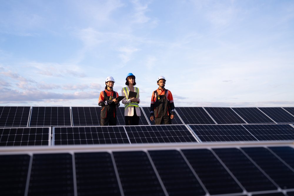 Three engineers wearing safety helmets inspect solar panels arranged outdoors under an overcast sky at a solar power plant, Madrid, Spain.