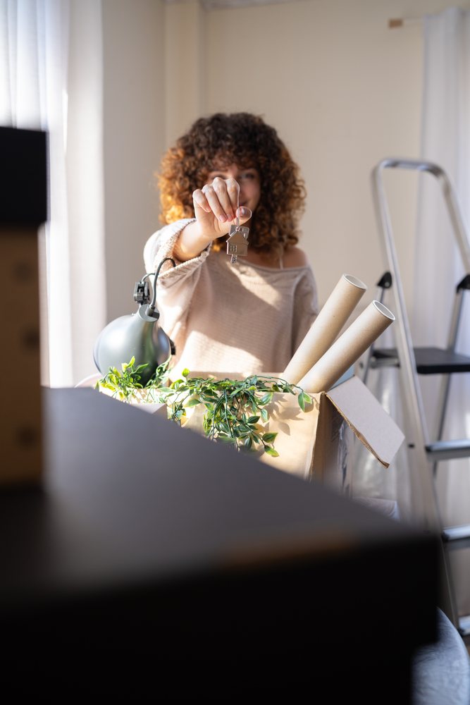 Woman holds a house key indoors near packed moving boxes containing a green plant, lamp, and cardboard rolls, Spain.