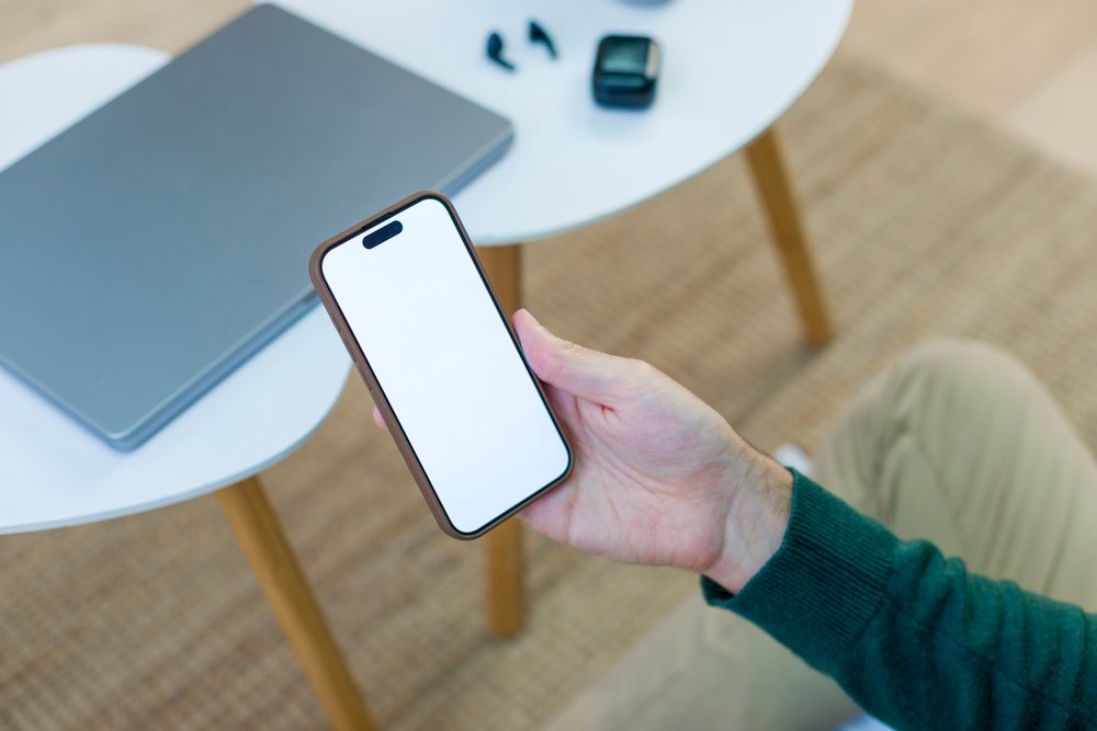 Person holding a smartphone with a blank screen over a white table in a modern home office with a laptop and car key fob visible, technology, young adult.