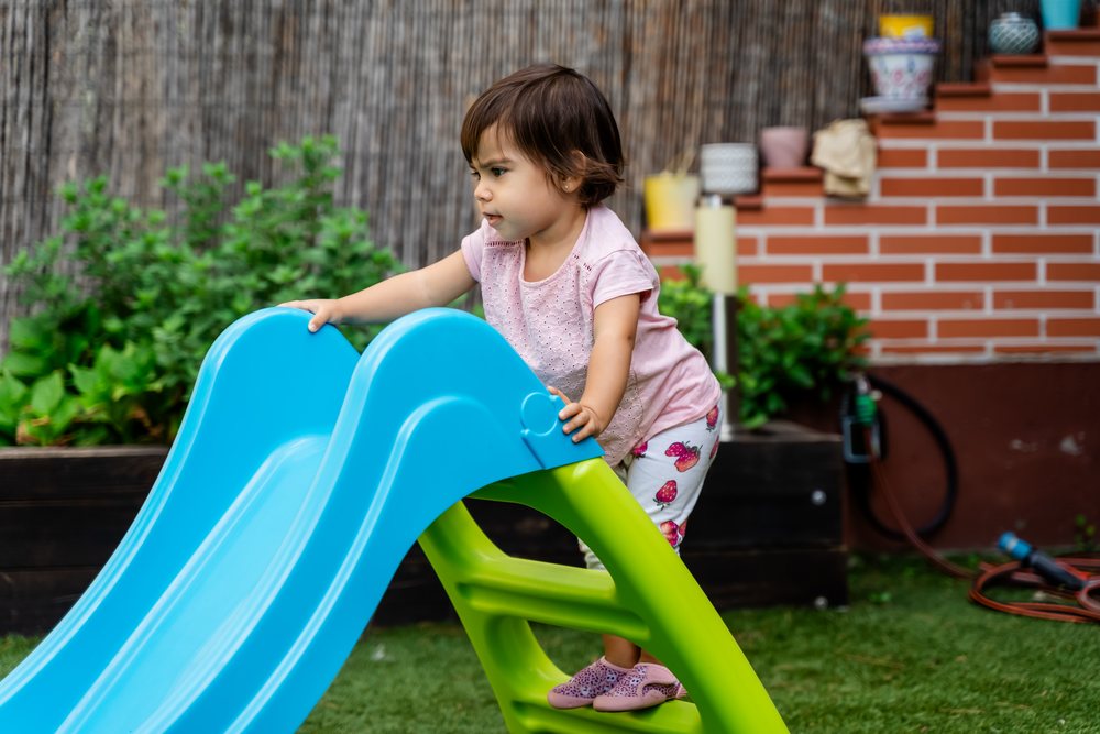 Toddler wearing pink shirt and strawberry print pants climbs a blue and green plastic slide in a backyard garden with green plants and a brick wall, children, Spain.