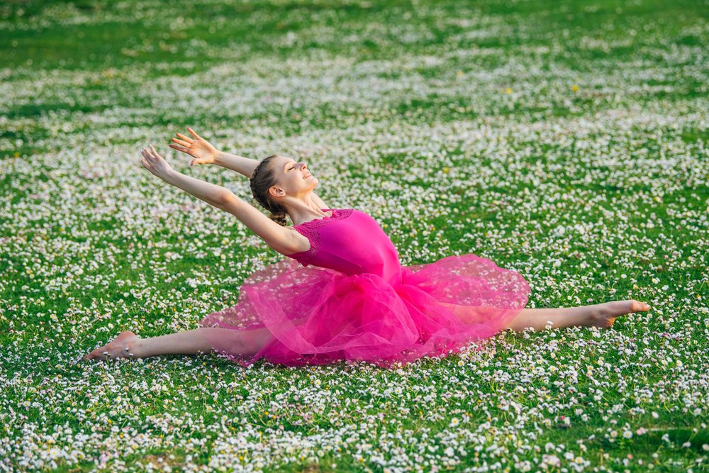Female ballet dancer wearing pink tutu and leotard performing a split stretch outdoors on a green grass field covered with white daisies in daylight, USA.