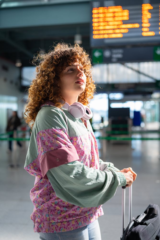 Young woman with curly hair and headphones waits in a train station holding luggage under daylight, Spain.