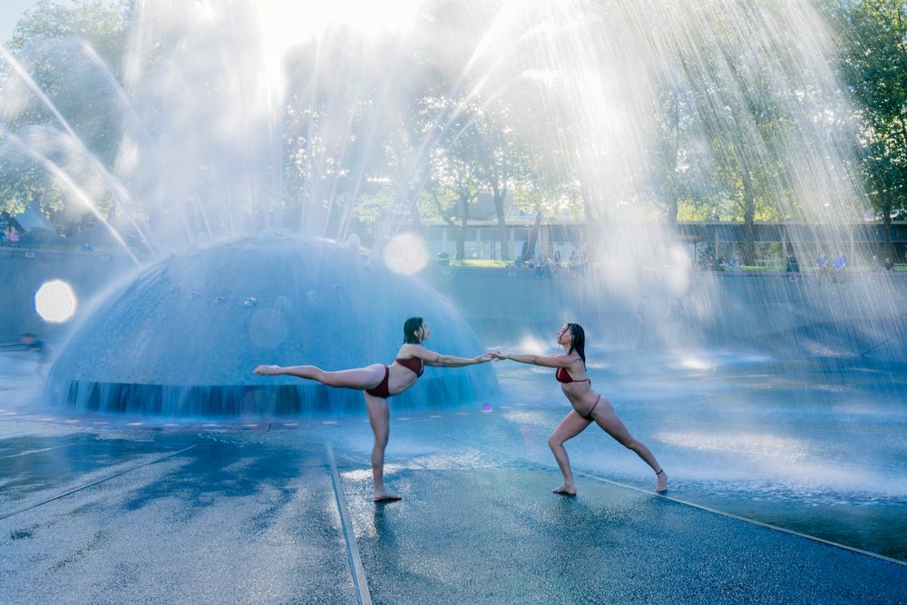 Two women performing partner acrobatics yoga poses by an outdoor water fountain with water sprays, USA.