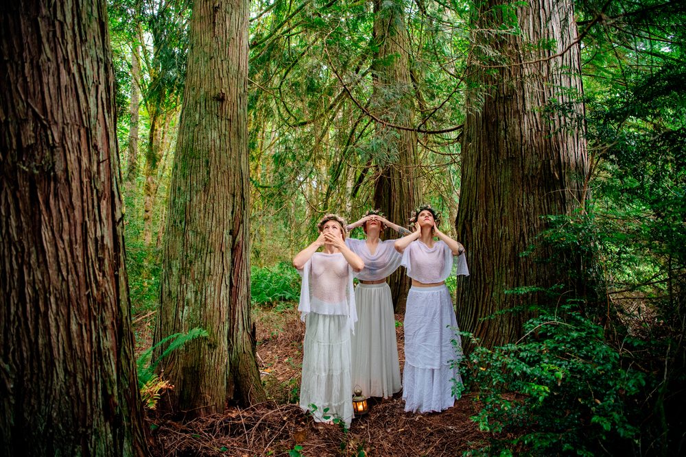 Three women wearing white clothing and flower crowns stand barefoot in a dense forest in the USA with a lantern on the ground, muses.