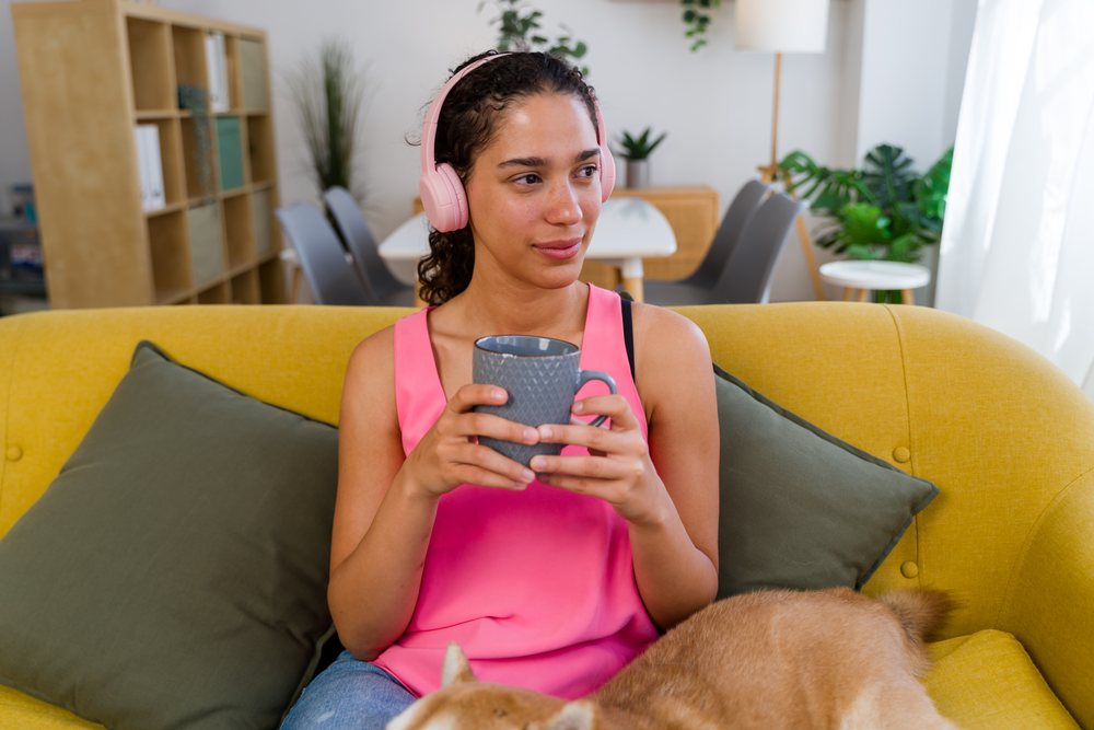 A woman wearing pink headphones and holding a mug relaxes on a yellow couch with a dog in a modern living room, Spain.