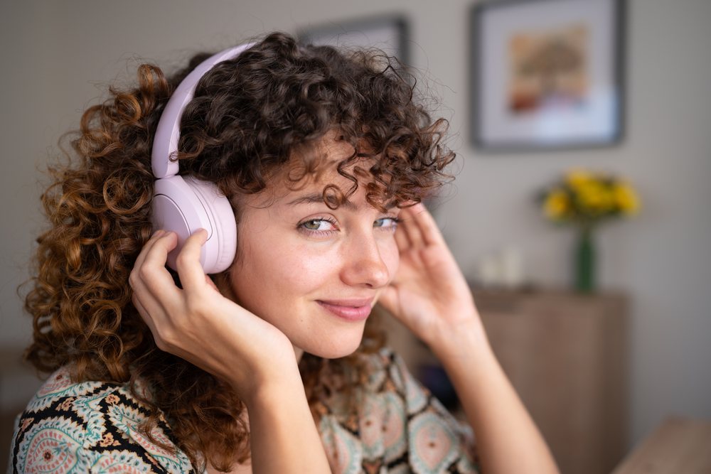 Woman with curly hair smiles while listening to music on pink headphones indoors at home with yellow flowers in the background, Spain.
