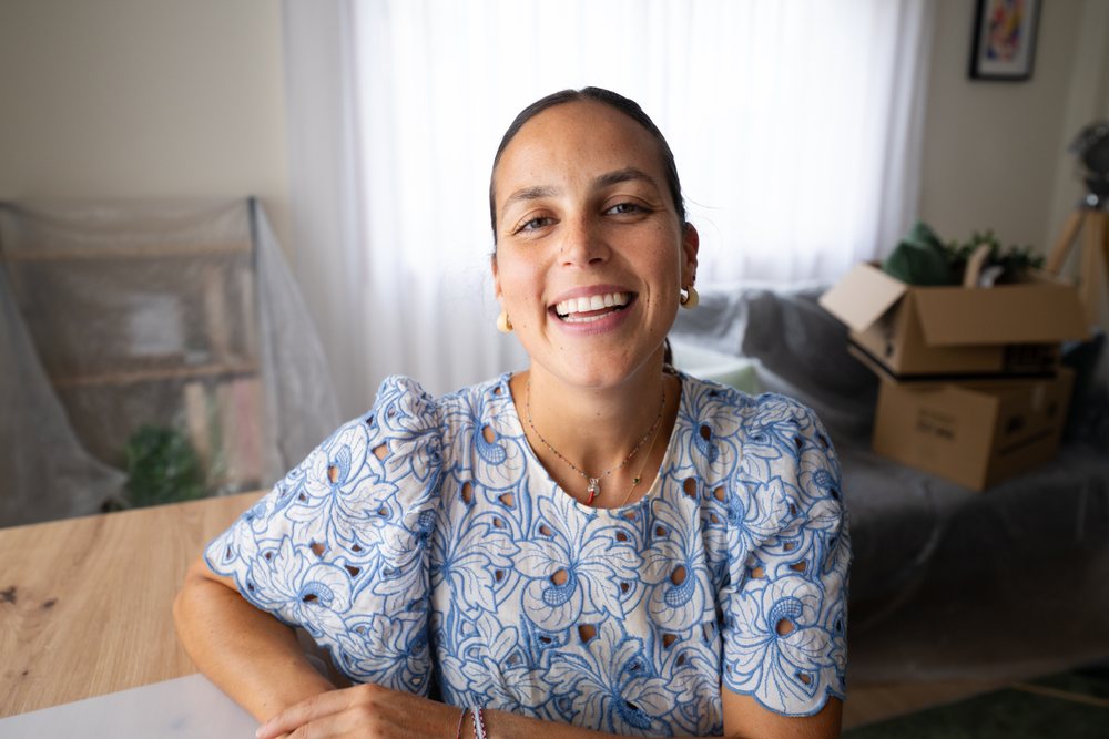 Smiling woman wearing earrings and a necklace sits at a wooden table in a home office with natural light and cardboard boxes stacked on a covered couch, Spain.