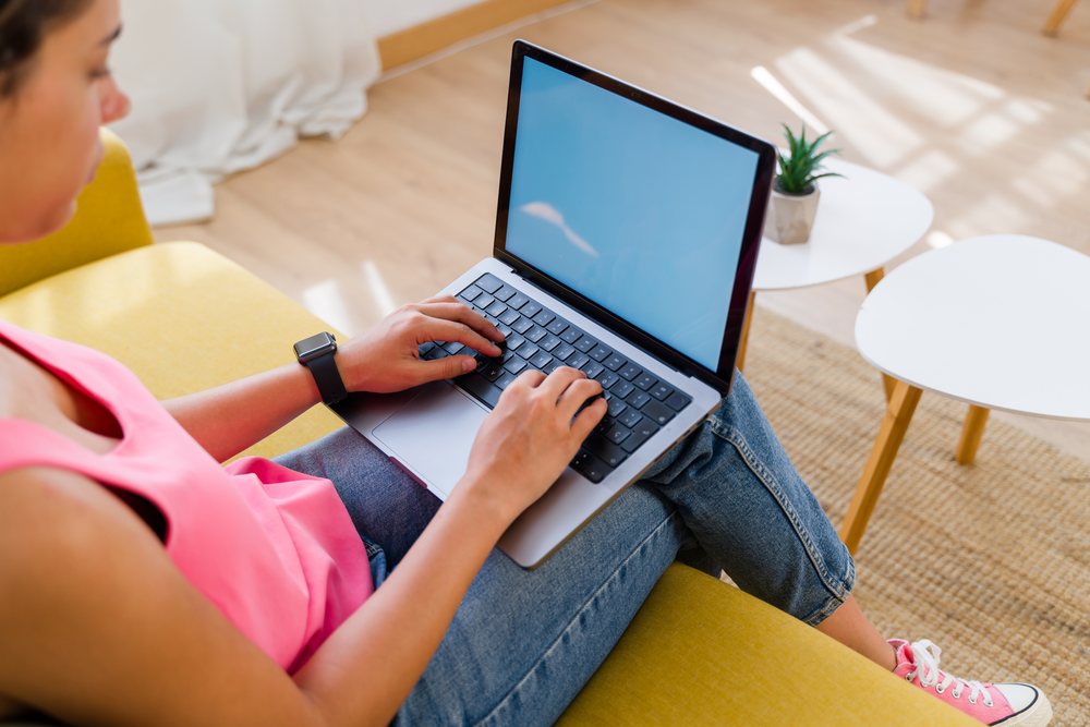 A woman is typing on a laptop while sitting on a yellow sofa in a modern, naturally lit living room, Spain.