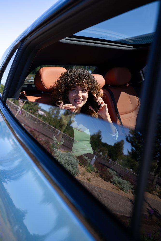 Woman with curly hair talks on a mobile phone inside a car with brown leather seats and sunroof, reflecting the road outside, remote work, businesswoman, Spain.