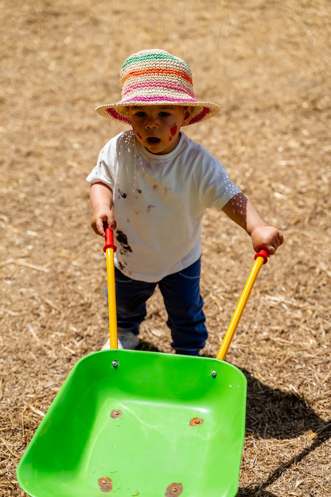 Child wearing a straw hat and face paint pushing a green toy wheelbarrow on straw-covered ground outdoors in daylight, kids, Spain.
