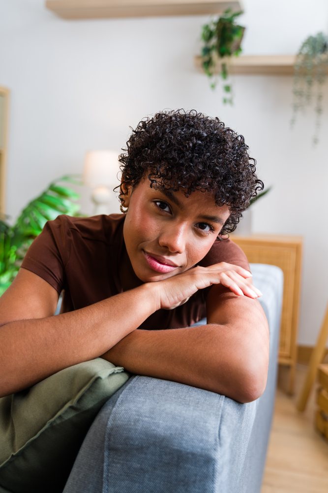 Woman with curly hair resting her arms on a grey sofa in a cozy indoor setting with green plants and soft lighting, Spain.