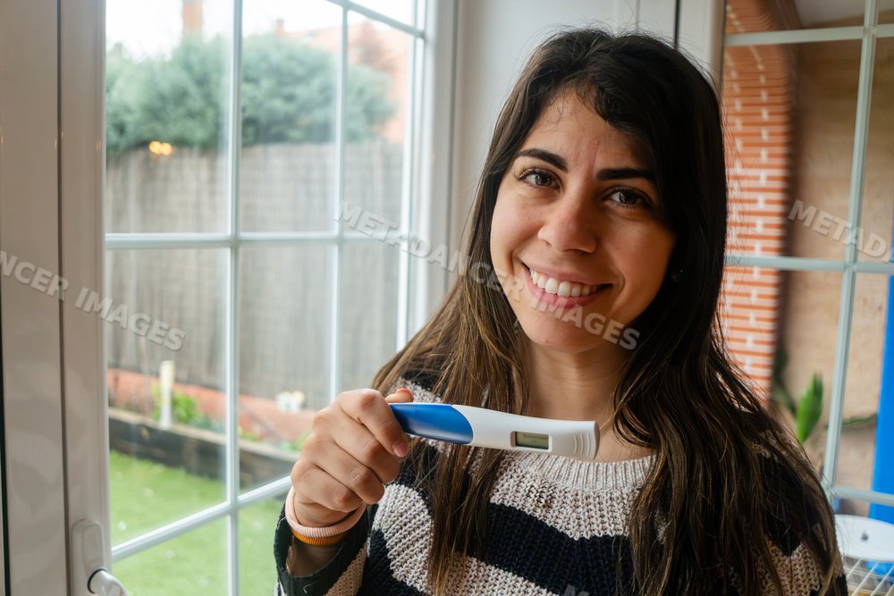 A smiling white woman holds a digital pregnancy test indoors