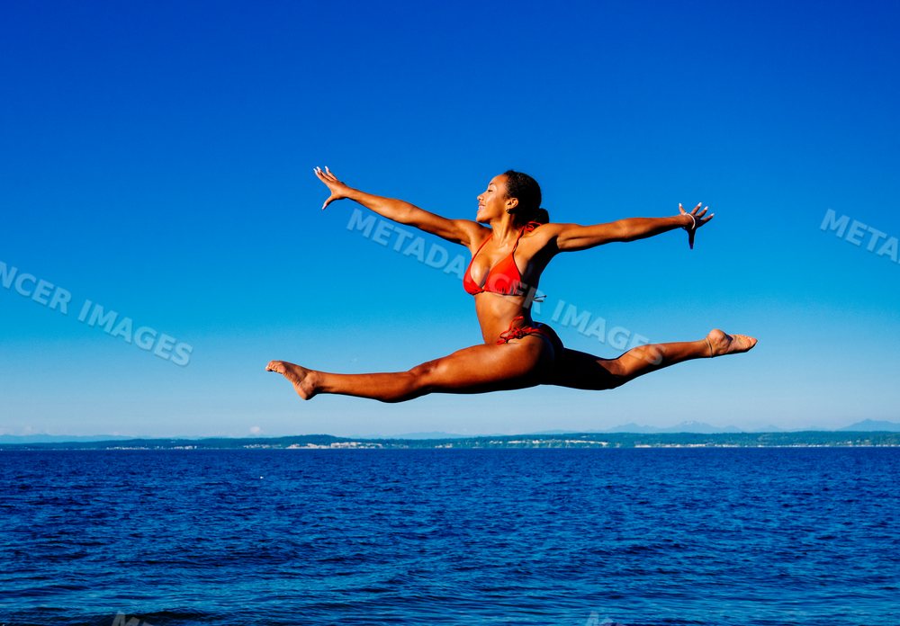 A woman in a red bikini jumps in a split over the ocean in WA, USA