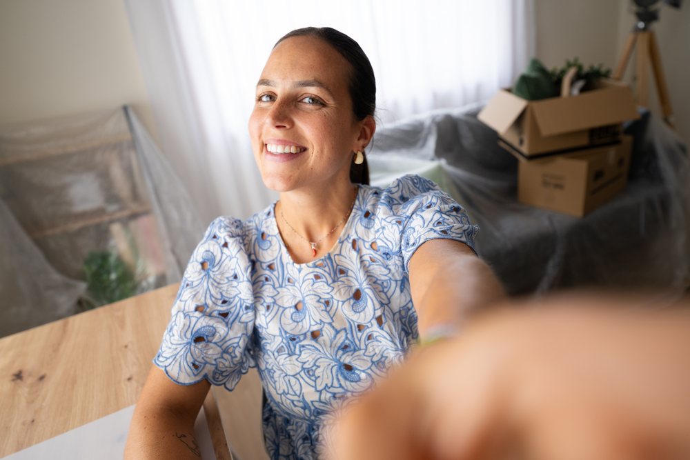 Woman smiles while taking a selfie indoors near covered furniture and stacked cardboard boxes in a home setting, telecommuting, Spain.