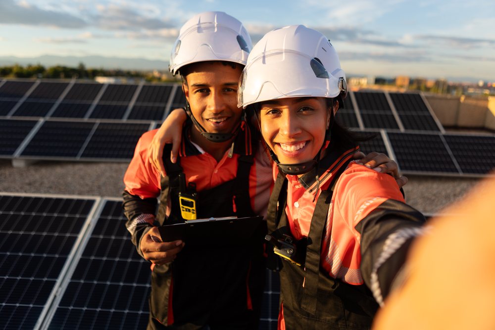 Two technicians wearing safety helmets take a selfie on a rooftop solar panel installation in sunlight, Madrid, Spain.