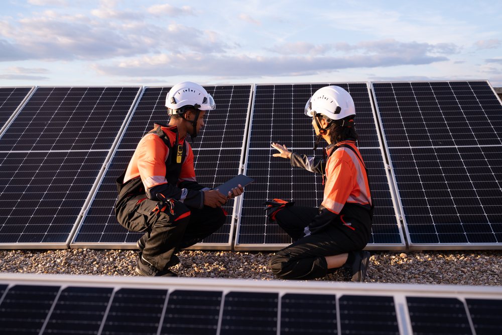 Two workers wearing protective helmets and reflective clothing inspect solar panels outdoors on gravel ground during sunset, Madrid, Spain.