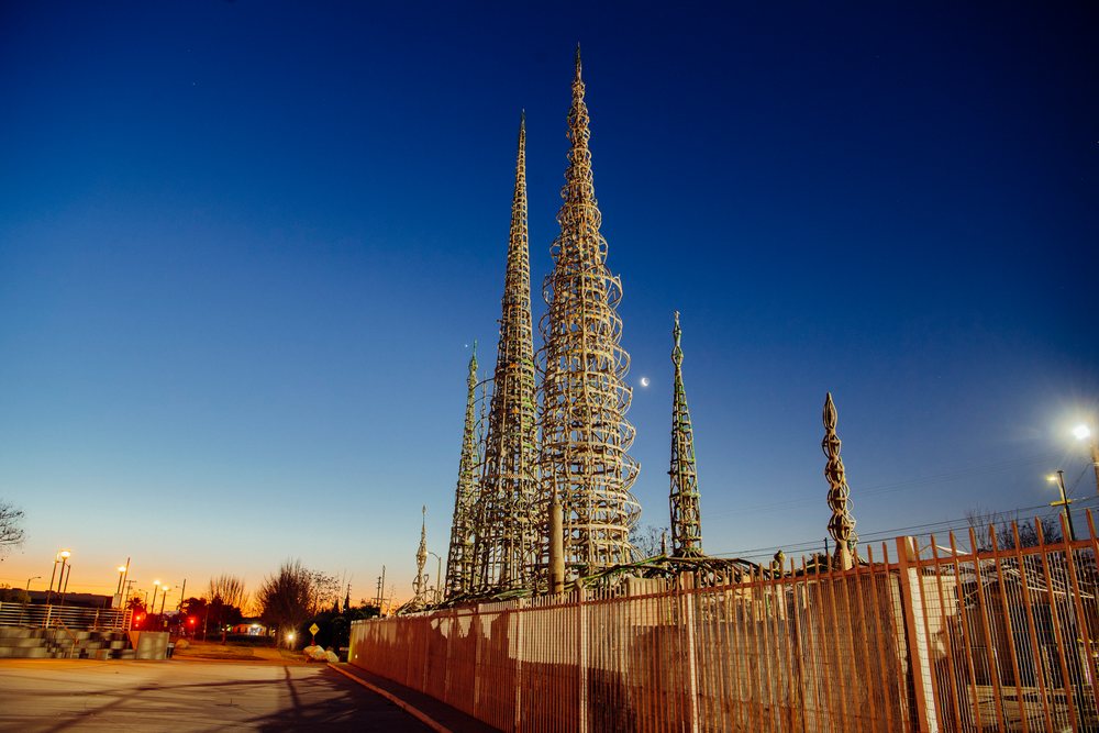 Steel and wire Watts Towers sculpture at twilight behind a fence in an urban outdoor California setting with crescent moon and street lighting, USA.