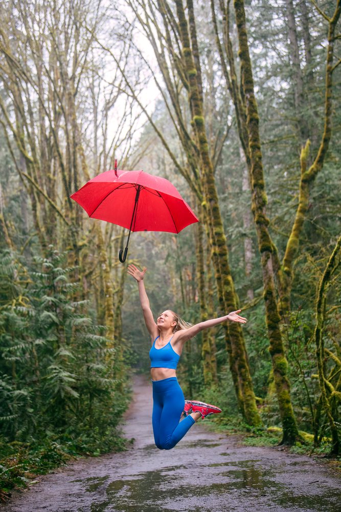 Woman in blue athletic wear jumps on a wet forest trail holding a red umbrella amid mossy trees and rain droplets, USA.