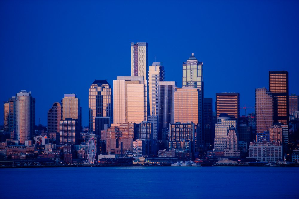 Seattle city skyline at dusk showing waterfront with a large ferris wheel and reflections on the water under a clear blue sky, USA.