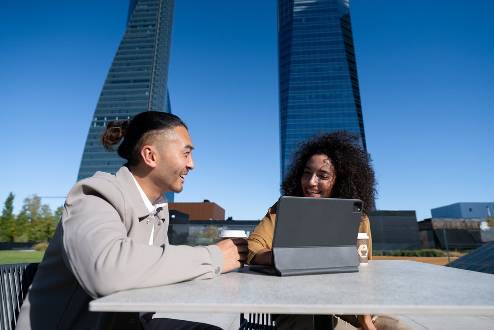 Two adults smiling and talking at an outdoor urban cafe table with a tablet and coffee cups, Madrid, Spain.