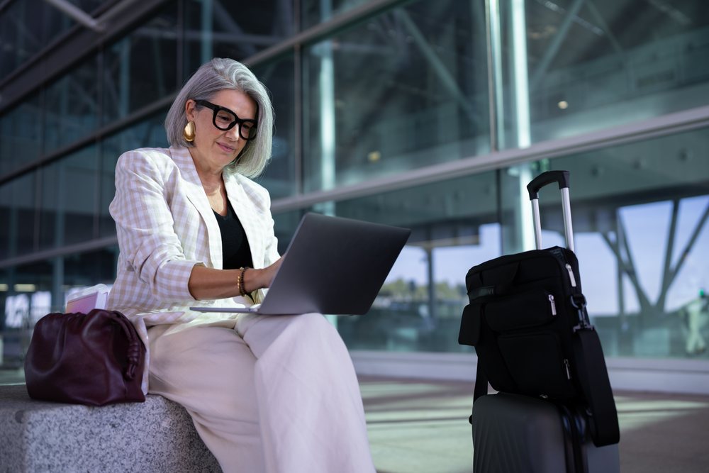 Mature businesswoman with gray hair works on a laptop outdoors at an airport, seated near luggage and travel bags, Spain, Madrid.