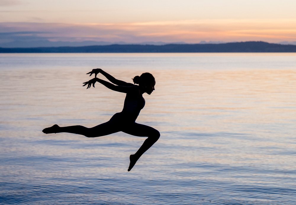 Silhouette of a woman performing a yoga jump pose by a lake at sunset, USA.