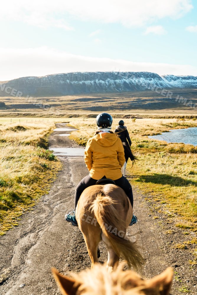 Two white people horseback riding on a trail beside a lake