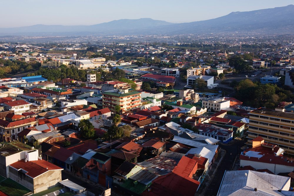 Daytime aerial view of San Jose, showing urban rooftops, scattered trees, roads, and a distant mountain range under clear sky, Costa Rica.