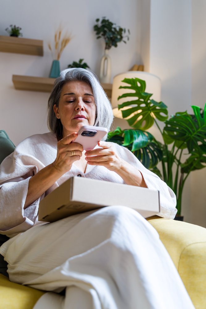 Senior woman with gray hair sits on a yellow chair indoors, holding a mobile phone with a cardboard box on her lap in a modern room, Spain.