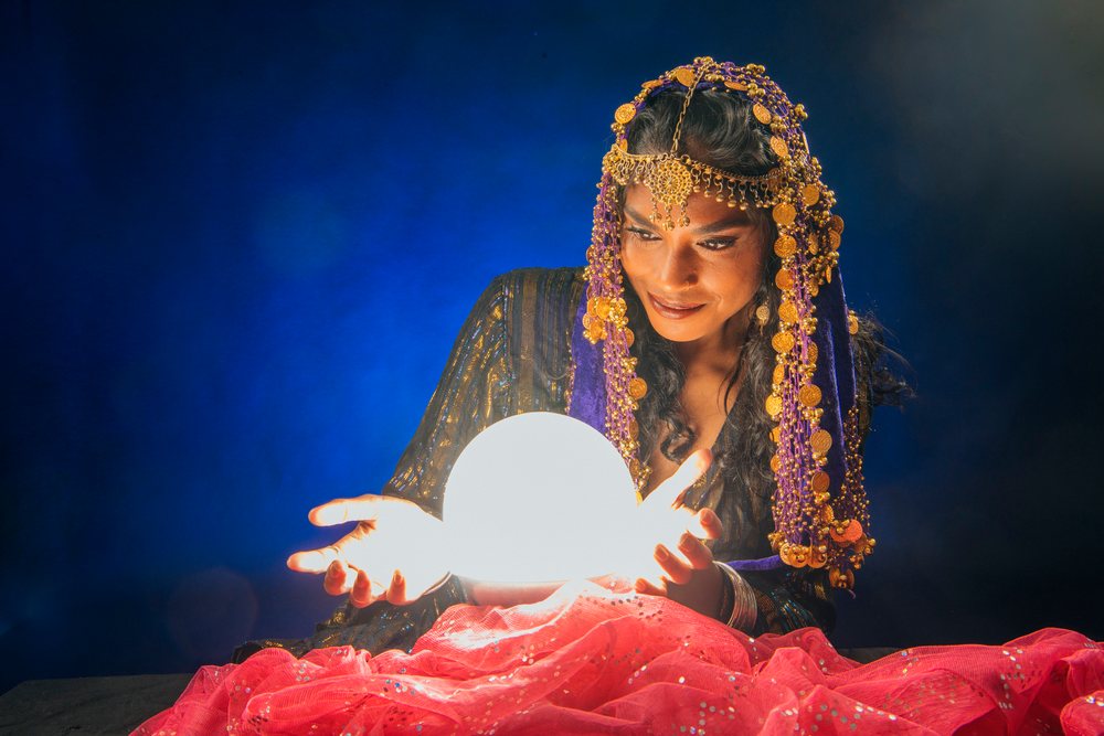 A woman wearing a purple headscarf adorned with gold coins gazes at a glowing crystal ball during a fortune telling session indoors in the, fortune teller, spirituality, USA.