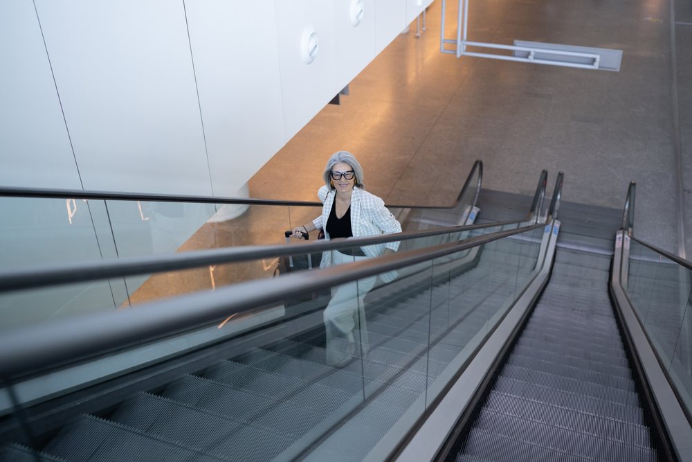 Businesswoman with gray hair and spectacles smiles while walking up an escalator carrying luggage inside a modern building, Spain, Madrid.