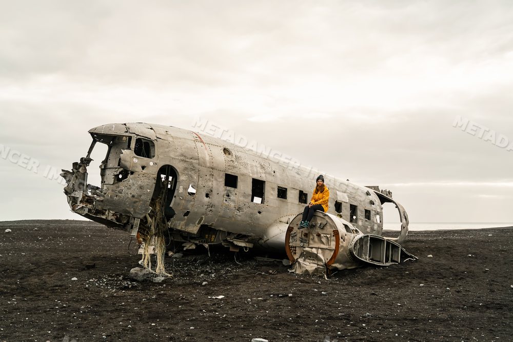 A white woman sits on wreckage of a crashed airplane on a black sand