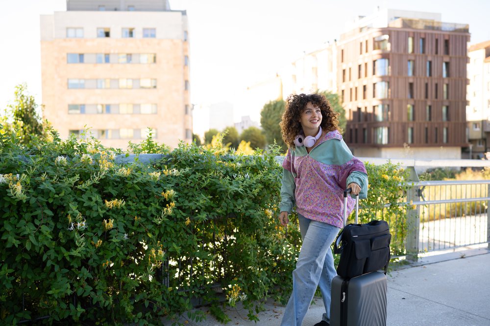Woman with curly hair and headphones walks with luggage along an urban sidewalk beside greenery and modern buildings in daylight, Spain.