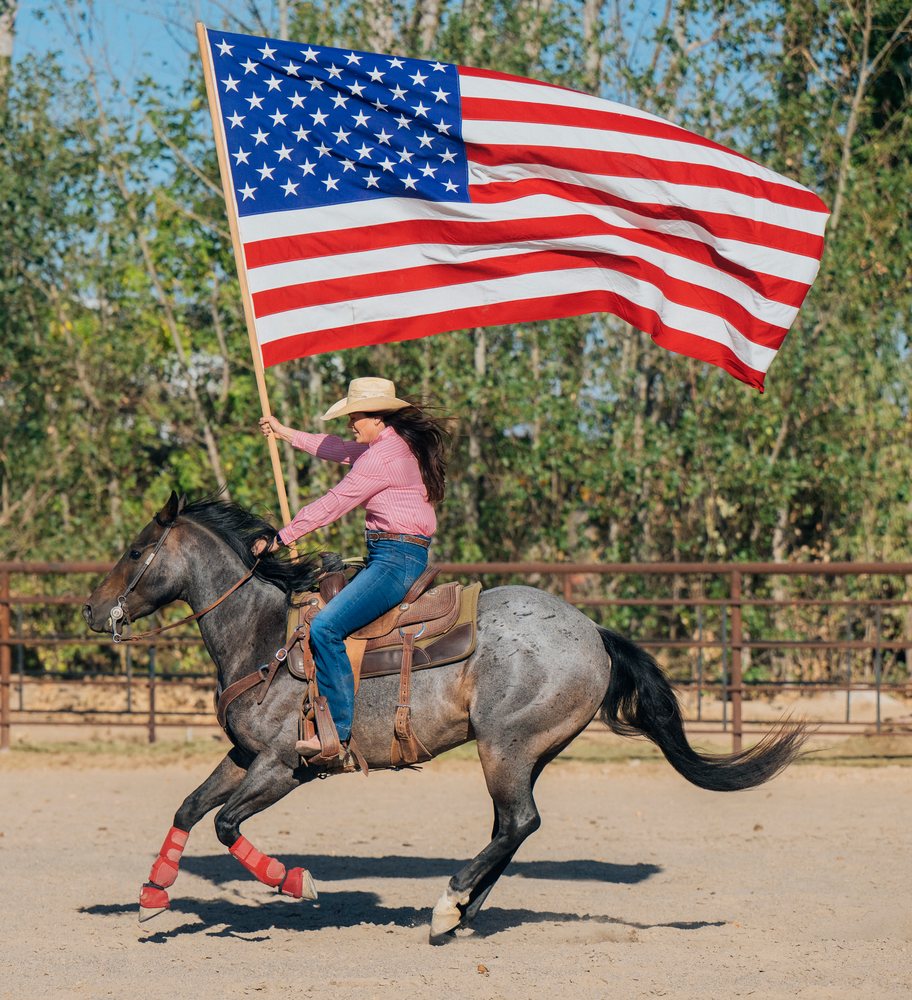 Person galloping on a saddled horse in an outdoor fenced arena in the Southwest USA holding a large American flag.