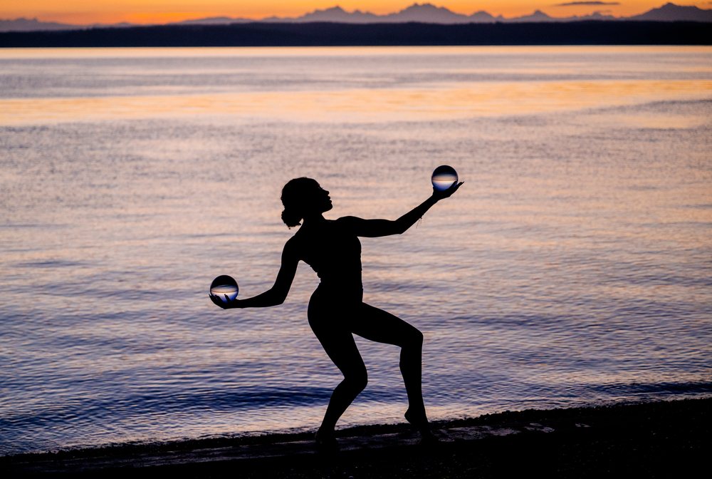 Woman in silhouette holding two glass balls while balancing in an acrobatic pose by a beach at sunset, yoga, acrobatics, USA.