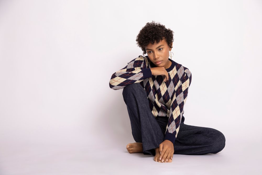 Young woman with curly hair sitting barefoot on the floor in a studio with a white background, wearing an argyle sweater and long pants, looking thoughtful, fashion, young culture.