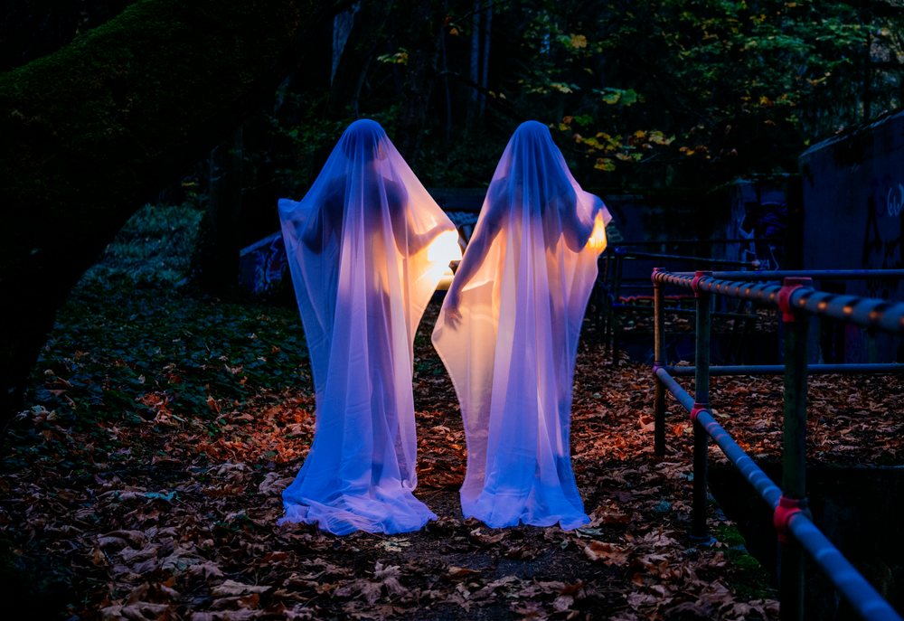 Two ghost figures draped in veils hold glowing lights on a forest path covered with fallen leaves at night, ghosts, USA.