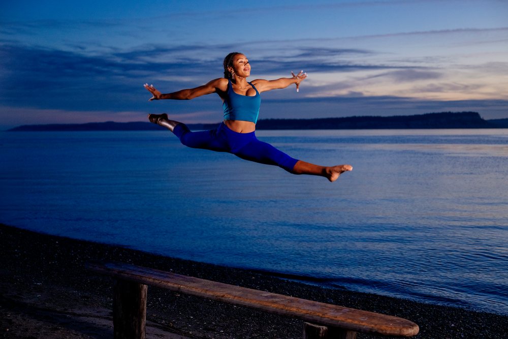 Woman wearing blue workout clothes leaps in an acrobatic pose along a beach at sunset, yoga, acrobatics, USA.